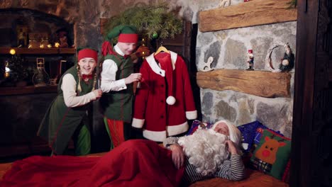 santa claus is sleeping in his bedroom at christmas time. funny elves are waking him with a ringing bell. christmas and new year concept