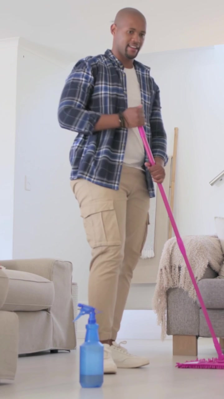 Vertikal video: En mann i rutete skjorte plasserer en rosa mopp, sprayer fra en blå flaske og mopper stuegulvet for å rengjøre.