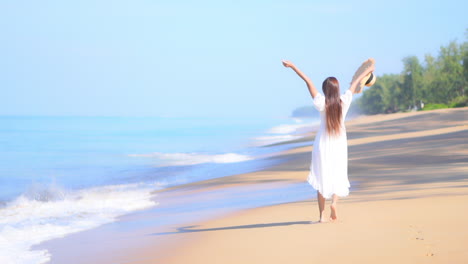 a happy young woman, in a white flowing sundress carrying a large sun hat, dances along the beach as the waves roll in