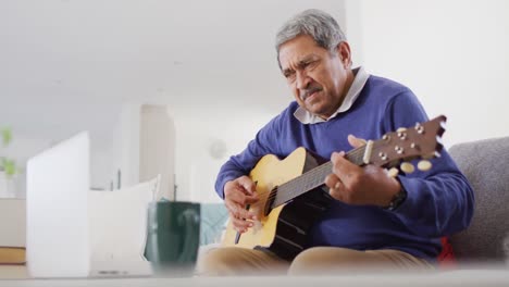 Video-Eines-Lächelnden-älteren-Gemischtrassigen-Mannes,-Der-Zu-Hause-Einen-Laptop-Benutzt-Und-Akustikgitarre-Spielt