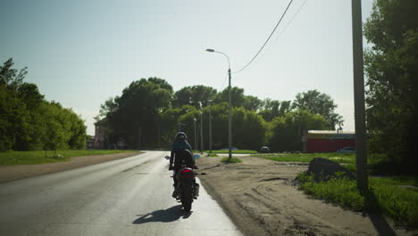 a female biker navigates a serene road on her power bike, the road is flanked by trees, electric poles, and parked cars, with buildings visible in the background