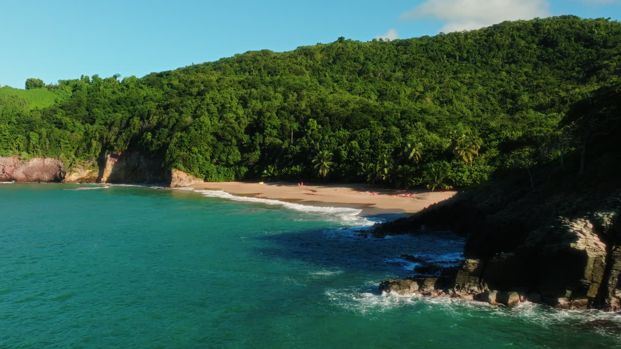 FPV drone flies fast along the rocky coast of Guadeloupe, descending toward turquoise water and moving toward a sandy tropical beach