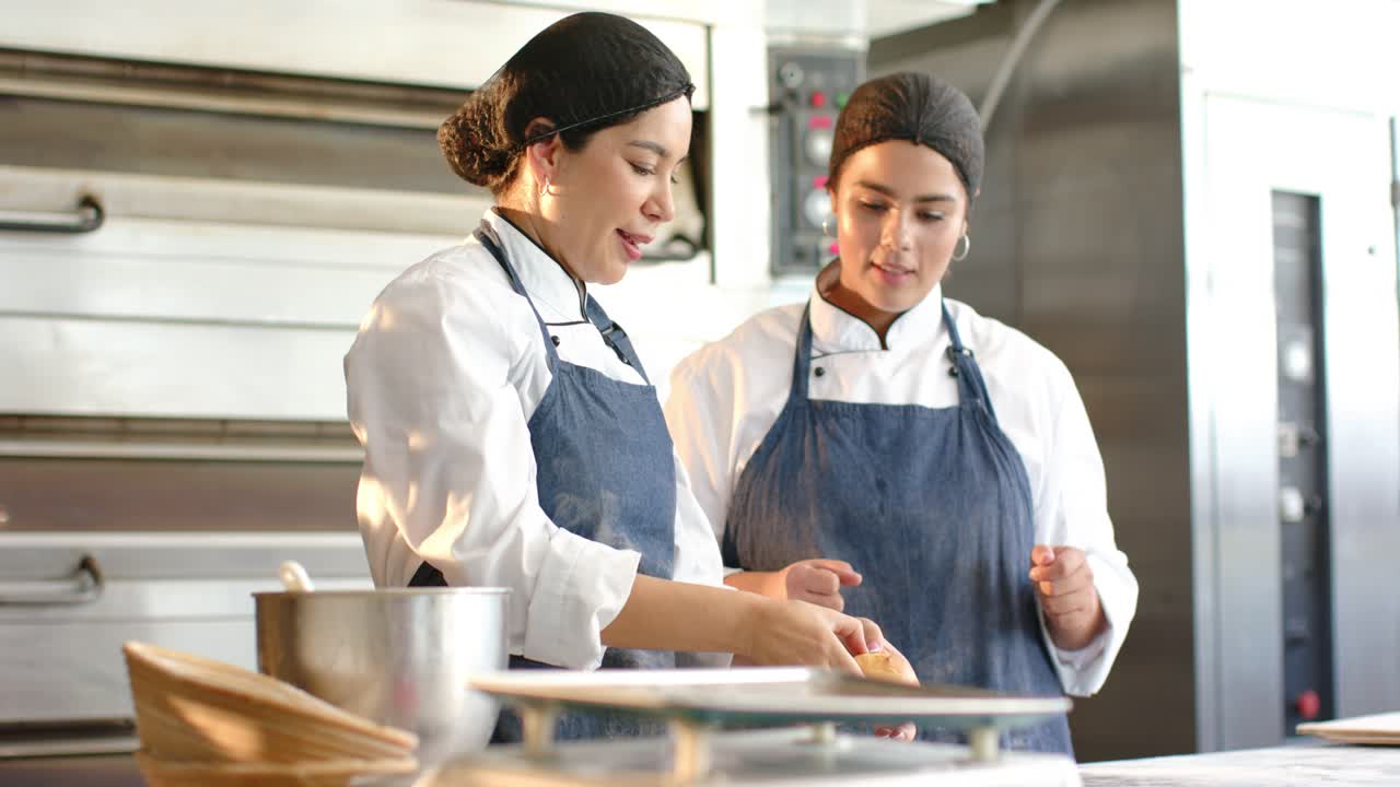 Diversas panaderas con chaquetas de chef en un mostrador de preparación, una mentora selecciona un bollo mientras enseña a una aprendiz.