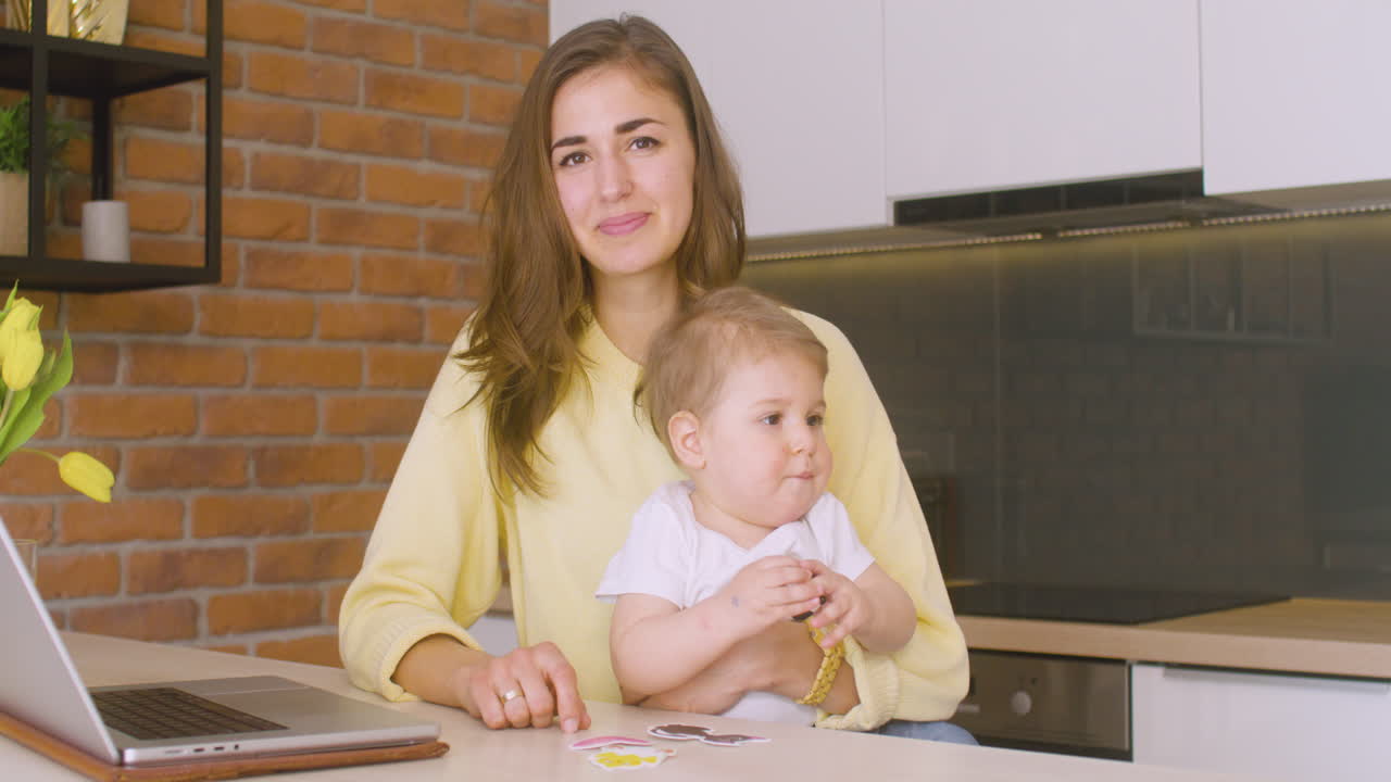 mujer mirando a la cámara mientras está sentada en la cocina y sosteniendo a su bebé en su regazo