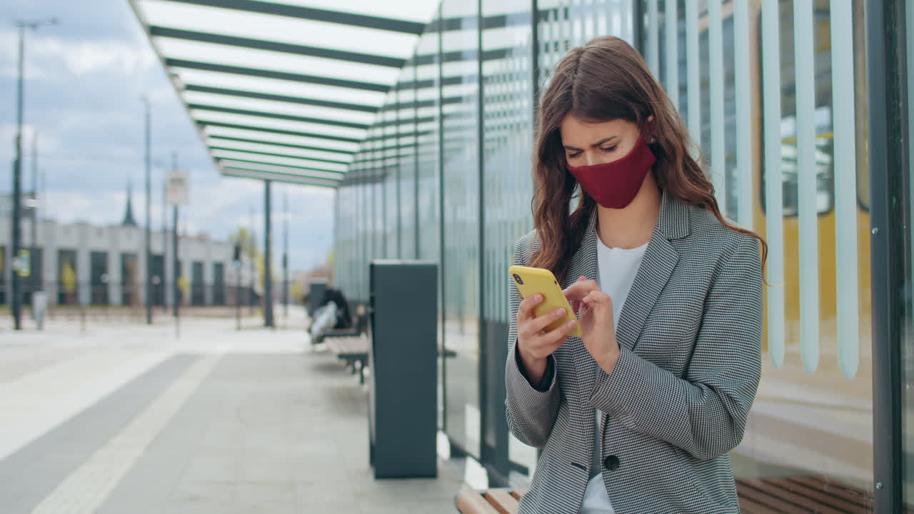 Young woman in a face mask using her smartphone at a modern public transport stop