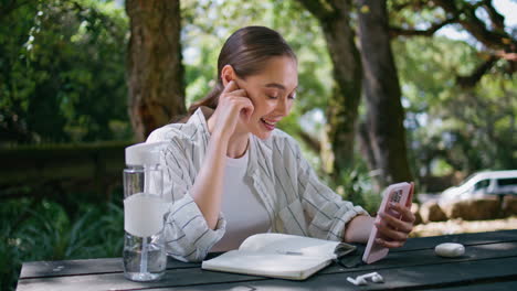 cheerful student calling online smartphone sitting with notebook at table park.