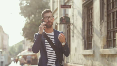 Hombre-Guapo-Con-Ropa-Elegante-Y-Gafas-Hablando-Por-Teléfono-Móvil-Mientras-Camina-Por-El-Casco-Antiguo