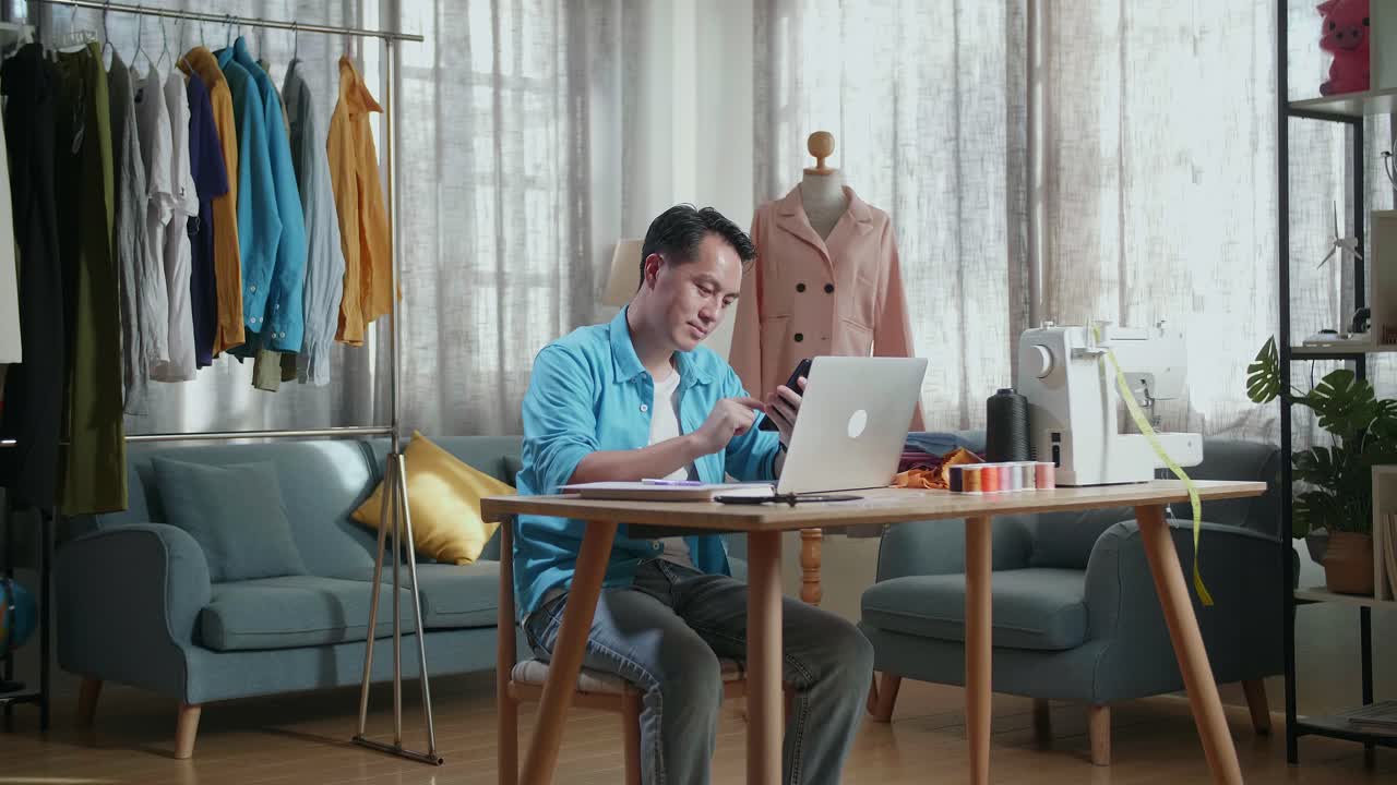 Asian Male Designer With Sewing Machine Looking At Smartphone While Designing Clothes On Laptop Computer In The Studio