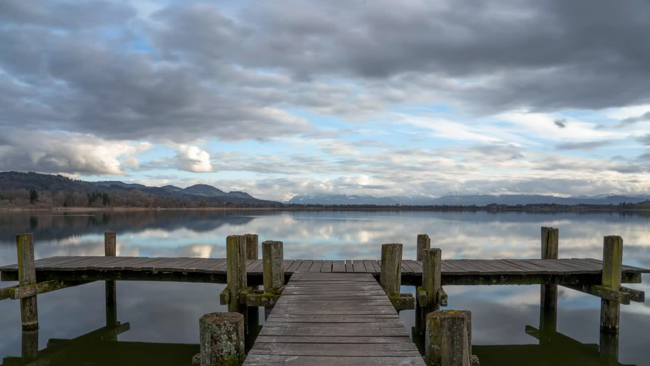 las nubes se reflejan en las aguas serenas del lago pfäffikersee suiza con un muelle de madera vacío y montañas impresionantes