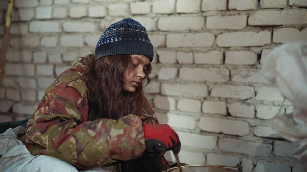 Survivor in camouflage jacket and knitted hat stirs pot with red gloves while companion hand reaches to drop leaf into food inside cold brick shelter, showing struggle for survival in harsh condition