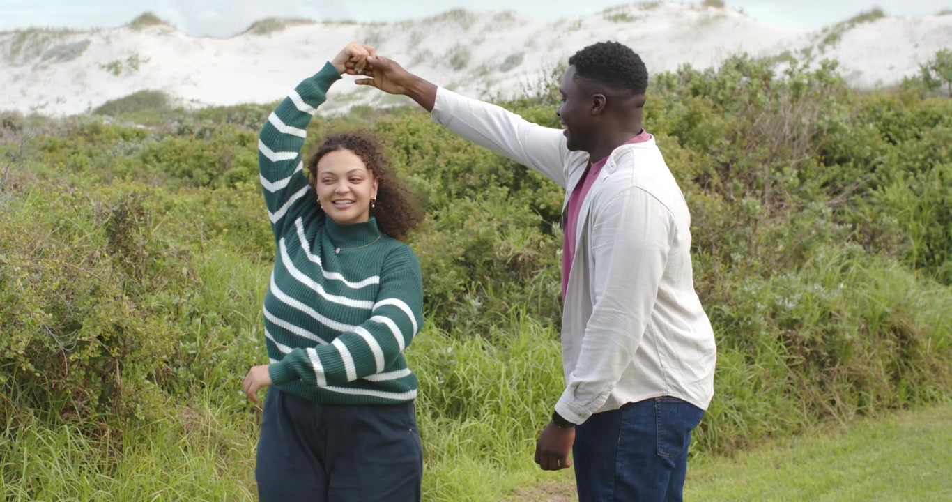 Afroamerikanisches Paar auf einem Dünenpfad; die Frau mit gestreiftem Oberteil bietet dem Mann die Hand, der sie zu einer Drehung führt, während sie ihre Liebe teilen.