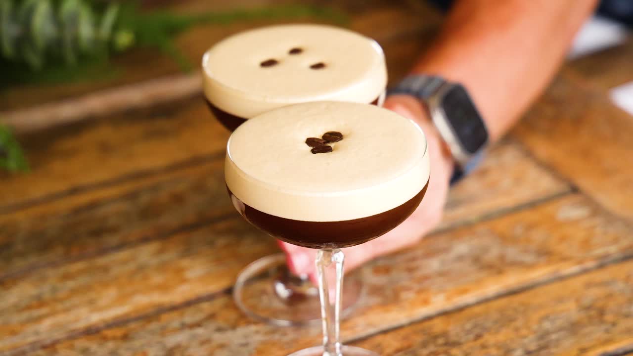 A hand places an espresso martini on a wooden bar counter. Bright lighting highlights the cocktail's creamy top
