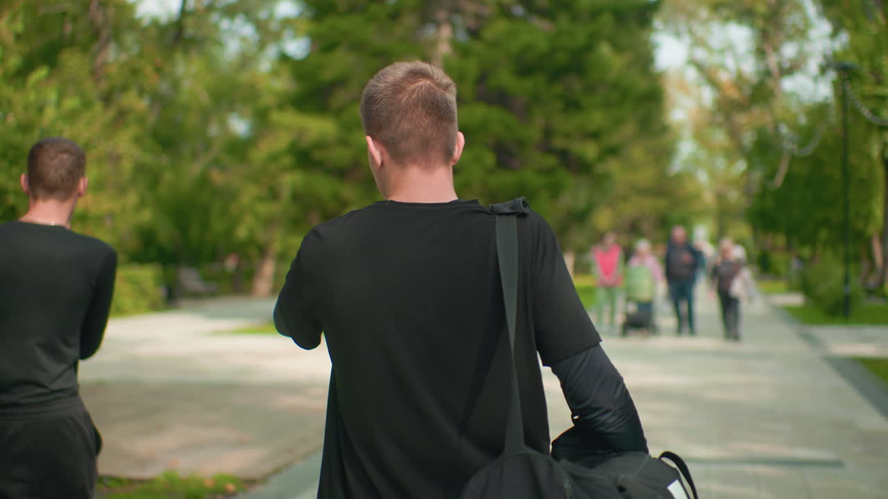 young white man walking down park path, carrying duffel bag toward distant pedestrians among sunlit trees, casual black outfit, steady pace, campus commuter energy, reflective mood and soft bokeh