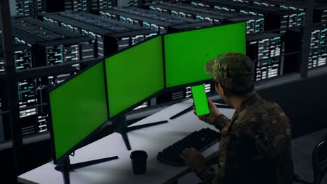 side view of happy asian military looking at mobile phone green screen and celebrating while working with multiple computer monitor in data center