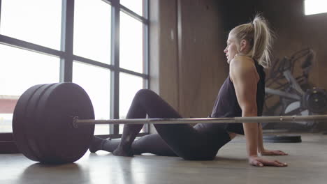 Out-of-breath-athletic-Swedish-woman-sits-on-floor-next-to-barbell,-gym-workout