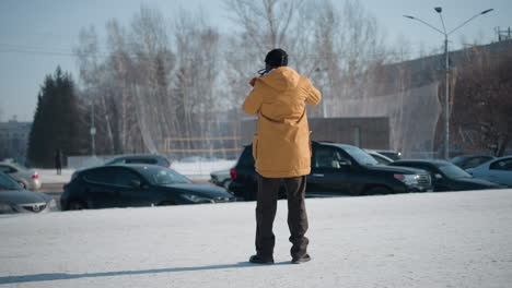 back view professional photographer in yellow winter coat capturing parked cars on snowy city plaza, gloves on, sturdy boots on snow, architecture traffic blur background under daylight glow
