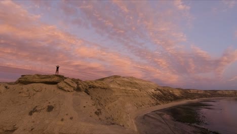silueta de hombre joven parado en una montaña con las manos detrás de la cabeza mirando al mar al atardecer - disparo de drones