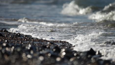 Low-close-up-shot-of-ocean-waves-rolling-up-on-a-pebble-covered-beach-in-dark-gray-and-brown-tones,-slow-motion