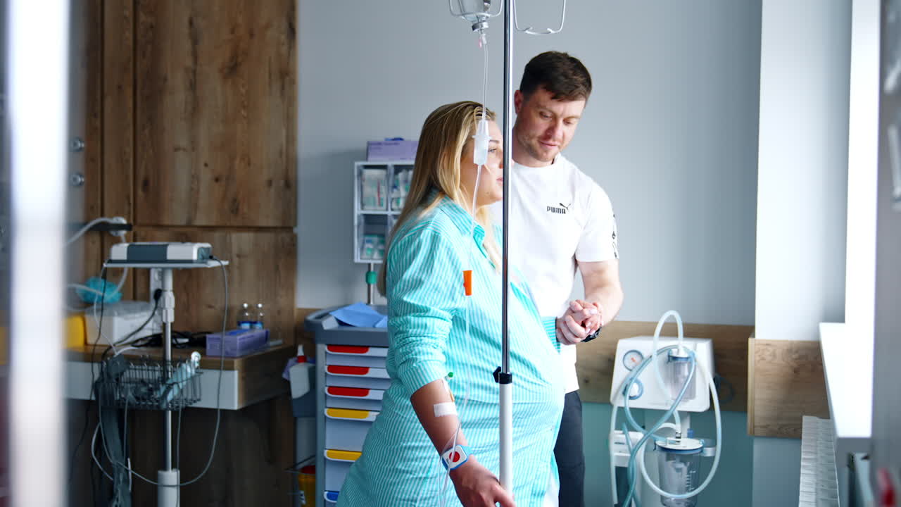 Blonde Caucasian woman expecting a child is in the hospital room. Pregnant lady walks supported by her husband and looks at window.