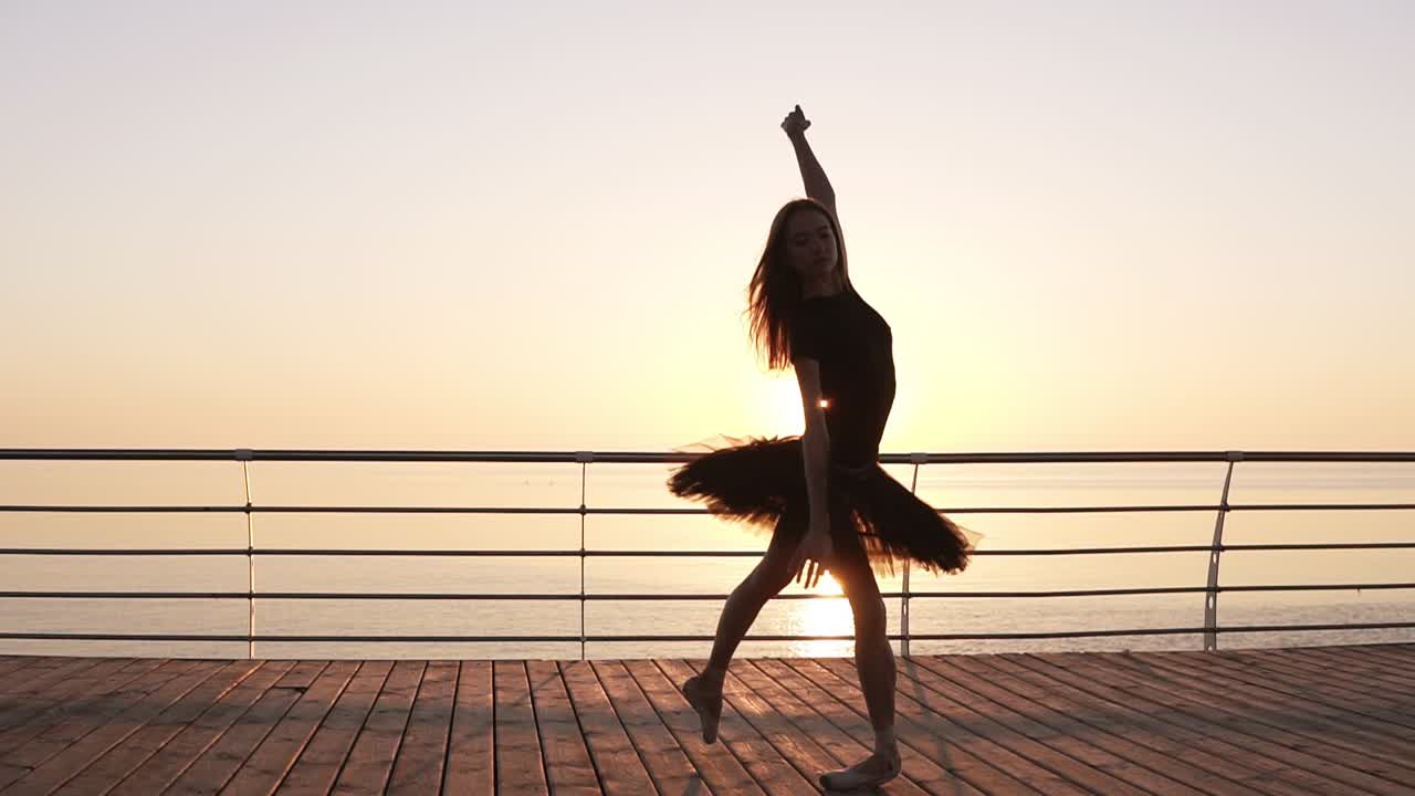 Dancing ballerina in black ballet tutu and pointe on seafront above ocean or sea. Morning bosk. Young attractive woman practicing, making vertical twine, lunge forward in the dance