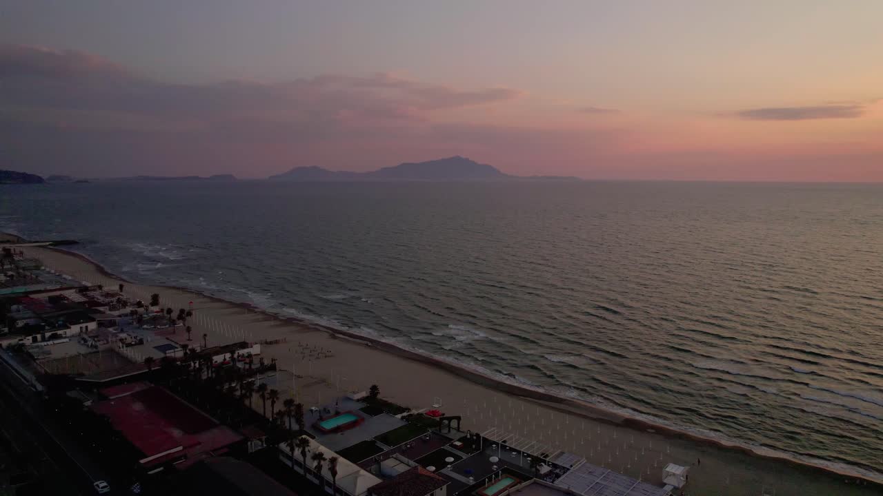 Drone captures quiet beach town along calm shoreline during sunrise. Soft waves roll in as pastel sky glows above distant mountains. Peaceful coastal scene in early morning light in Portugal