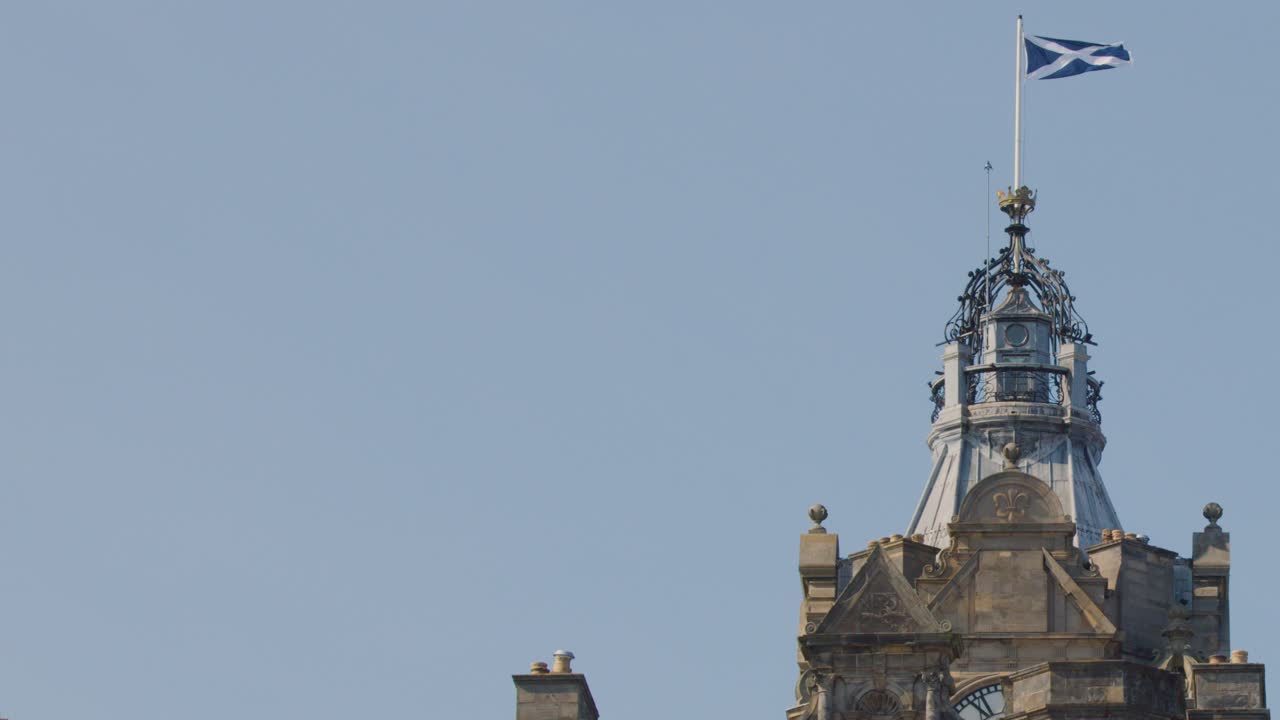 A static wide shot captures the Scottish flag fluttering atop a historic stone tower in Edinburgh under clear daylight, with minimal camera movement