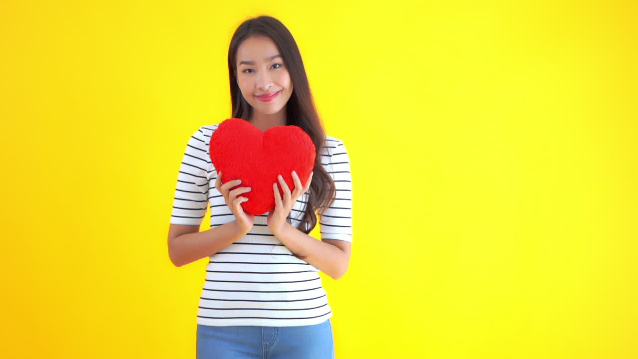 Asian woman in casual jeans and T-shirt showing fluffy red heart cushion smiling on a yellow studio background
