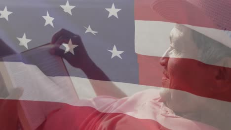 animation of flag of united states of america and senior biracial man reading book on beach