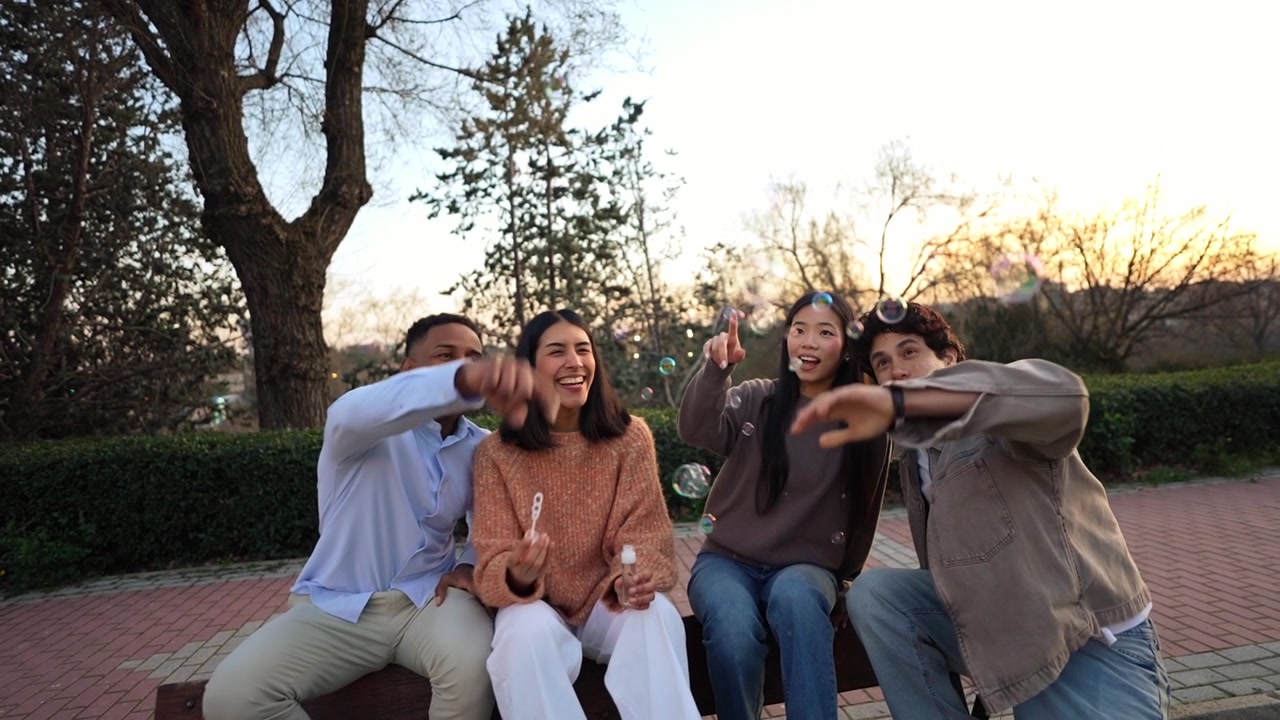 Amigos multiétnicos felices soplando burbujas y riendo en un banco del parque