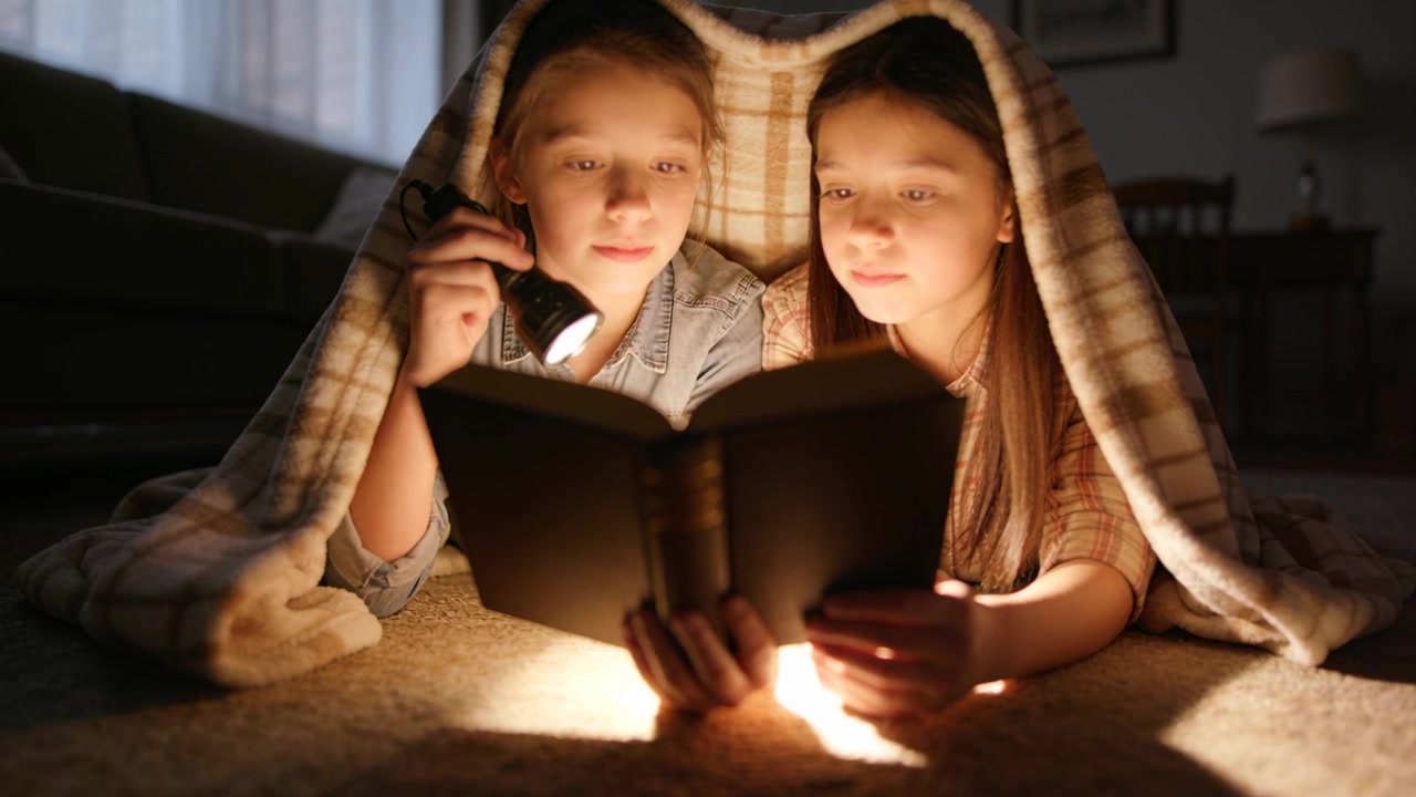Zwei Mädchen im Vorpubertätsalter schalten eine Taschenlampe ein und lesen ein Buch unter einer karierten Decke zu Hause, bekleidet.