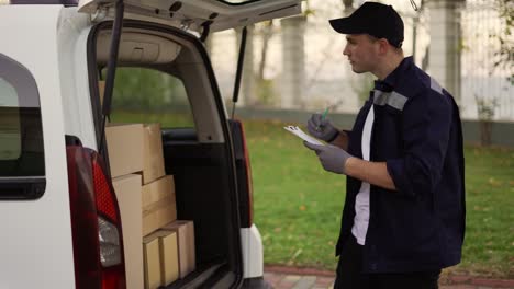 handsome courier making notes in documents and takes cardboard box package out of delivery car. courier in uniform, cap and