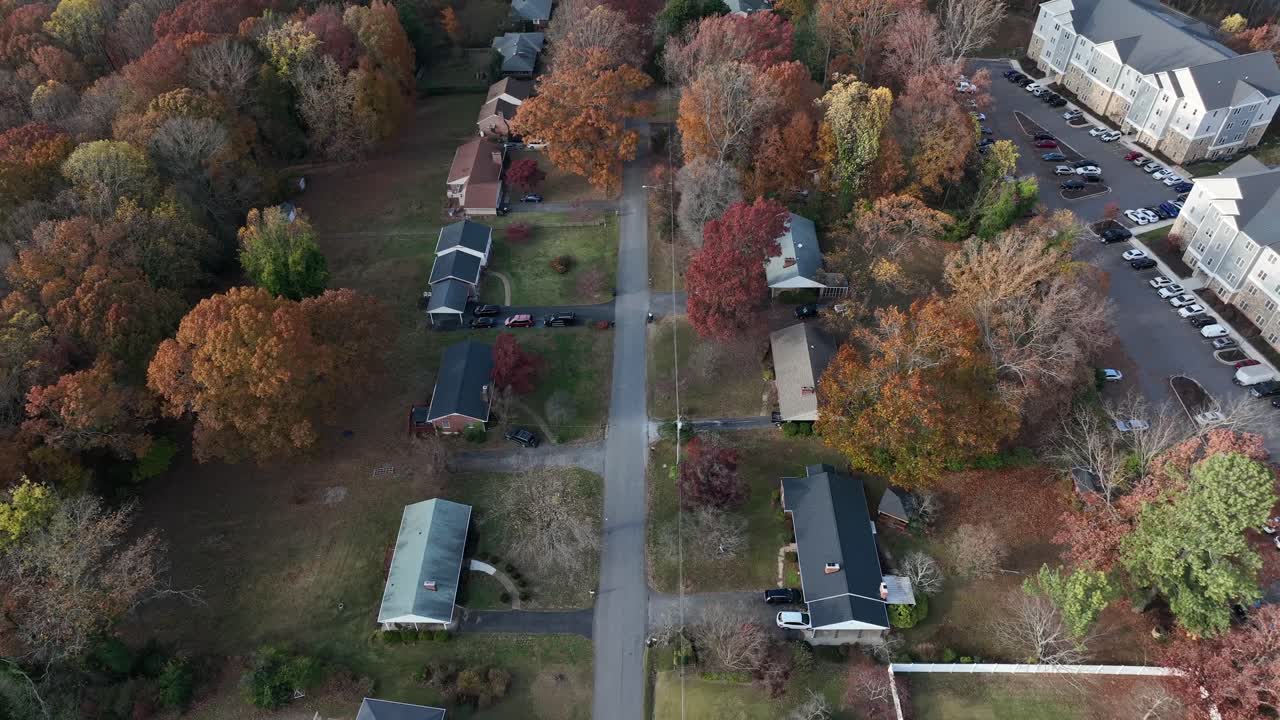 Aerial drone footage showcases an American suburban neighborhood in autumn, with a straight-line flight over a tree-lined street, colorful foliage, and houses under clear midday sunlight
