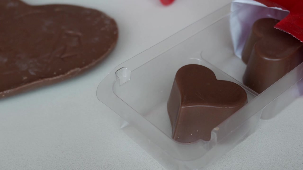Cioccolatini a forma di cuore in un vassoio trasparente accanto a una rosa decorativa su una superficie bianca.
