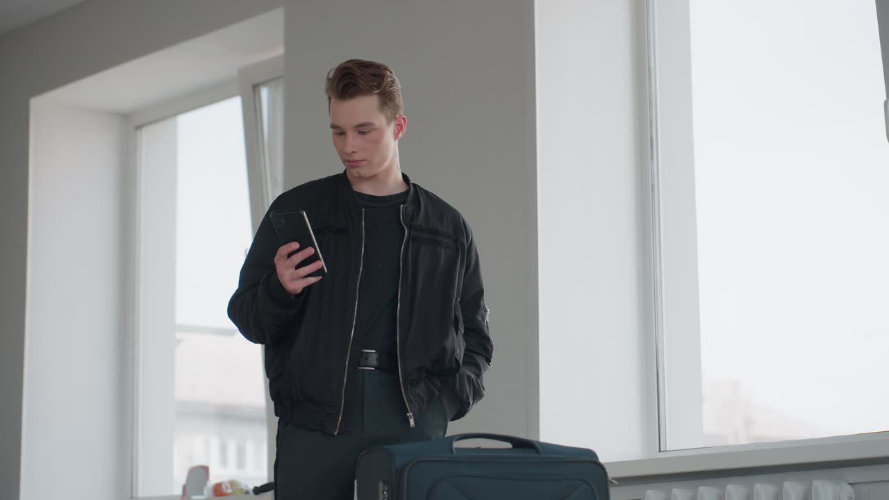 Boy in black jacket stands next to large wheeled bag, one hand in pocket while pressing smartphone against ear, bright window light and minimalist room