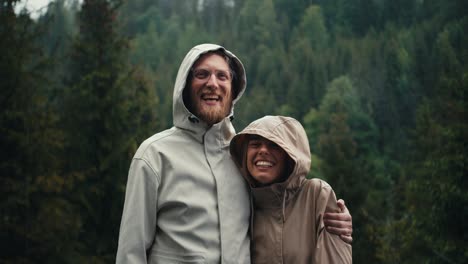 Happy-couple:-a-blond-guy-in-glasses-with-a-beard-and-a-blond-girl-in-jackets-stand-and-rejoice-against-the-backdrop-of-a-green-forest-in-the-mountains