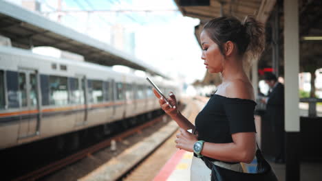 Una-Mujer-Con-Un-Top-Negro-Y-Pantalones-Blancos-Se-Encuentra-En-Una-Plataforma-De-Tren,-Mirando-Su-Teléfono-Inteligente,-El-Tren-Es-Visible-En-El-Fondo,-Lo-Que-Refleja-Una-Escena-De-Desplazamiento-Urbano