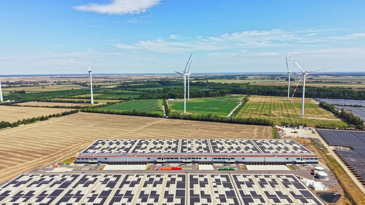 Wind turbines and solar panels seen near a large building in the countryside with fields and a blue sky in the background