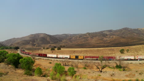 a freight train is parked along tracks below hells left burnt by wildfires - aerial parallax view