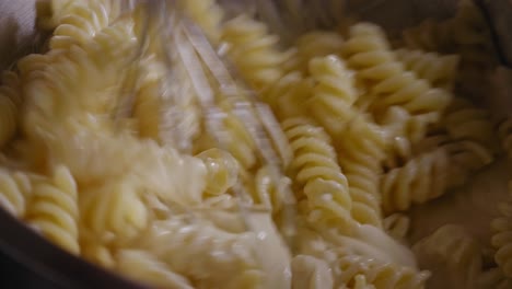 Vista-Cercana-De-Una-Mano-Usando-Un-Batidor-Revolviendo-En-Un-Tazón-De-Metal-Lleno-De-Pasta-Fusilli-Y-Masa-De-Queso-Crema---Disparo-Constante