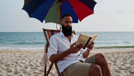 man reading a book on a beach chair under an umbrella