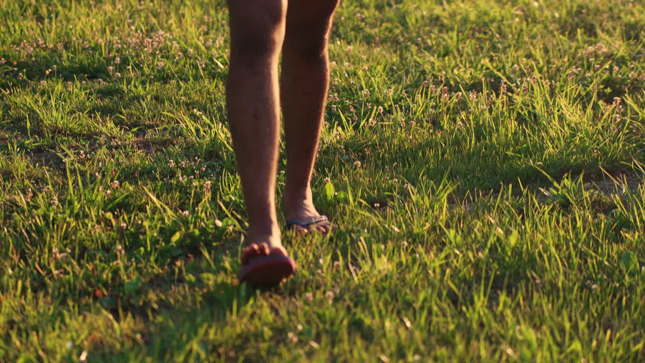 Tracking shot showing man legs wearing shorts and slippers and walking on lush green grass