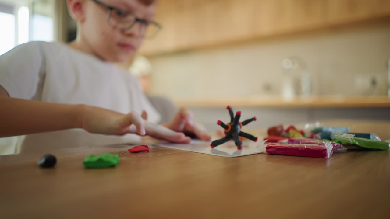 Child in white shirt sculpting black clay figure with red details at wooden table, surrounded by colorful packets of modeling clay, demonstrating creativity,and artistic play in bright home environment