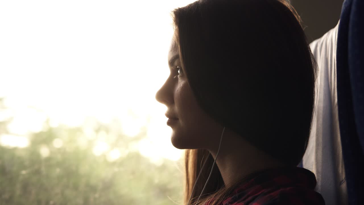 chica atractiva sentada en la sombra en el tren. escuchando música con auriculares y teléfono móvil. rayos de sol en la ventana. paisaje natural afuera