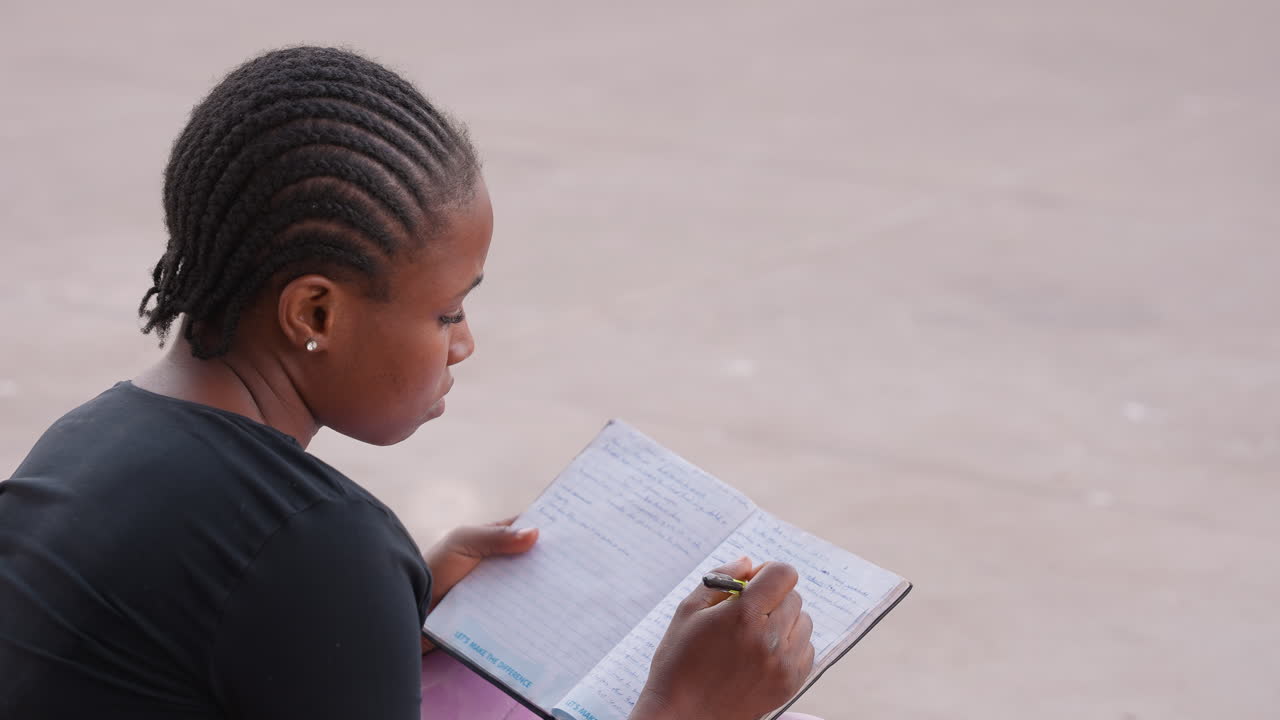 Ragazza che studia felicemente. Studentessa che scrive su un quaderno all'aperto. Donna concentrata sullo studio con i capelli intrecciati all'aperto. Giovane donna che prende diligentemente appunti su un quaderno durante una sessione di studio all'aperto.