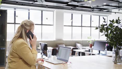 happy plus size caucasian casual businesswoman using laptop and talking on smartphone at desk