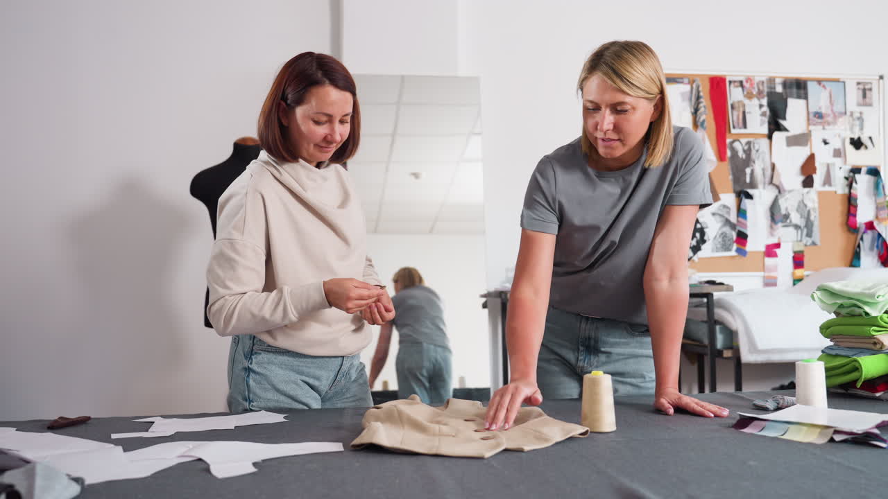 Garment designers in bright studio lean over beige jacket, discussing perfect spot to sew button, surrounded by pattern pieces, thread spools, fabric swatches, mannequin shadow