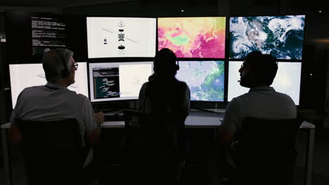 equipo de personas que trabajan en una sala de control con múltiples pantallas de computadora que muestran datos y mapas