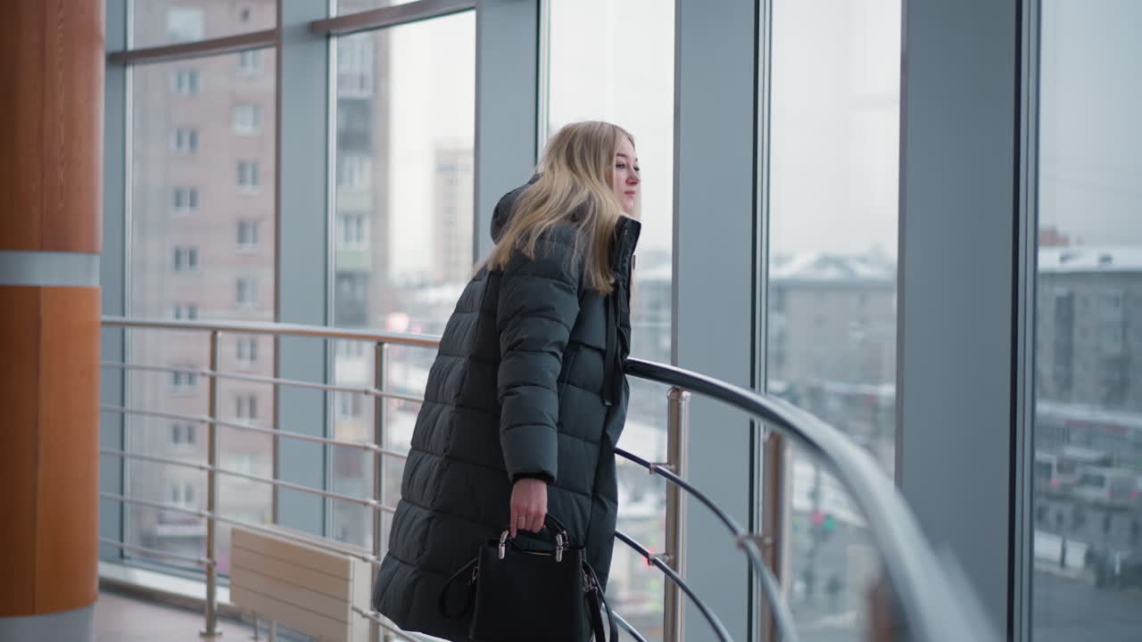 Lady standing by iron railing, looking out through glass with black handbag, cityscape in background, creating an atmosphere of contemplation and elegance in modern urban environment