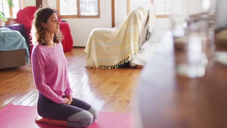 Mujer-Relajada-De-Raza-Mixta-Practicando-Yoga,-Arrodillada-Sobre-Una-Alfombra-Con-Los-Ojos-Cerrados-En-Un-Soleado-Dormitorio-De-Cabaña