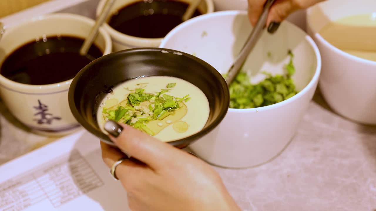 Hand uses tongs to add green herbs to creamy hotpot sauce at self-service condiment station